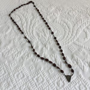 May include: A long necklace with dark red oval beads and a silver-colored pendant. The beads are strung together with a dark metal chain. The pendant is triangular with a detailed design. The necklace is displayed on a white, textured surface.