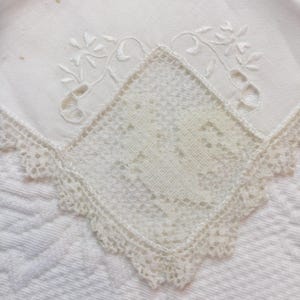 Puede incluir: Primer plano de un pañuelo blanco con delicado bordado floral y un centro de encaje en forma de diamante. Los bordes están acabados con un ribete de encaje festoneado, mostrando una estética vintage.