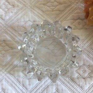 May include: A clear glass ring holder with a starburst design. The holder is made of glass and has a smooth, polished finish.