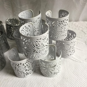 以下が含まれることがあります： 白い金属製のキャンドルホルダーのコレクション。複雑な花の切り抜きデザインが施されています。ホルダーは円筒形で、湾曲したハンドルが付いています。一部は積み重ねられ、その他は散らばっており、装飾的なディスプレイを作成しています。背景は白く、質感のある表面です。