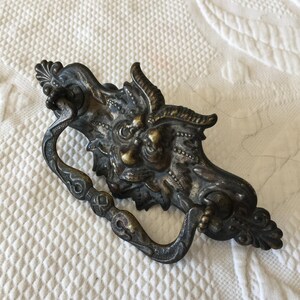 May include: A vintage ornate metal drawer pull with a detailed design of a mythical creature's face. The pull is made of a dark metal with a tarnished finish.