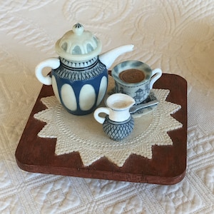 Op de afbeelding: Een miniatuur theeset op een houten basis met een witte kantonderlegger. De set bevat een theepot, een roomkan en een theekopje met schoteltje. De theepot en roomkan zijn blauw en wit met een bloemenmotief. Het theekopje is wit met een blauw en wit bloemenmotief.