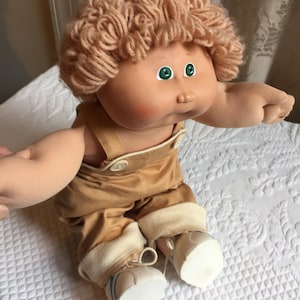 以下が含まれることがあります： ブロンドの巻き毛、緑色の目、バラ色の肌をしたキャベツパッチキッズ人形。人形は、白いトリムと白い靴が付いた茶色のオーバーオールを着ています。