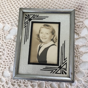 May include: A vintage silver-toned picture frame with a black and white photo of a smiling child. The frame has Art Deco-style black geometric corner designs. The photo is a portrait of a young girl wearing a dark vest and white collared shirt.