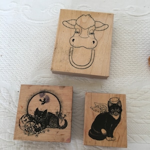 May include: Three wooden stamps with different designs. The first stamp features a black and white illustration of a cow's head. The second stamp features a black and white illustration of a cat with a halo and wings. The third stamp features a black and white illustration of a cat with a full moon in the background.