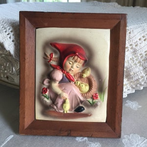 Może przedstawiać: Ceramiczna tabliczka ścienna w ramie przedstawiająca dziewczynkę w czerwonej sukience z kapturem i koszykiem piskląt. Tabliczka ma efekt 3D i jest pomalowana pastelowymi kolorami.