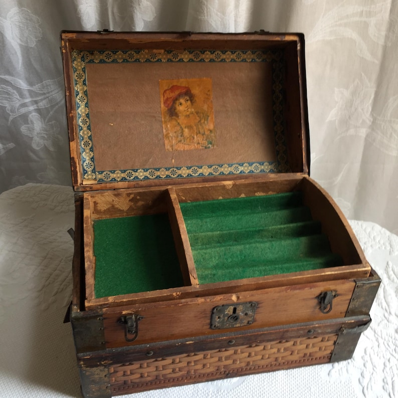 Puede incluir: Una caja de joyer&iacute;a de madera antigua con una tapa con bisagras y un patr&oacute;n tejido en la parte inferior. El interior est&aacute; forrado con fieltro verde y tiene un peque&ntilde;o compartimento para guardar joyas.