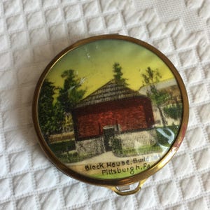 Op de afbeelding: Een ronde, gouden compact met een geschilderd beeld van een rood bakstenen gebouw met een geel dak. De tekst "Block House, Build 1764 Pittsburgh, Pa." staat gedrukt op de onderkant van de compact.