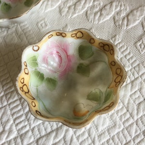 May include: A small, white porcelain bowl with a scalloped edge. The bowl is decorated with a hand-painted pink rose and green leaves. The rim of the bowl is trimmed with a gold band.