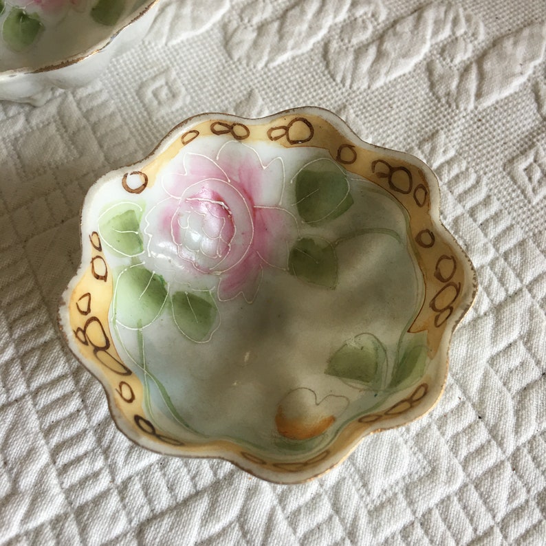 May include: A small, white porcelain bowl with a scalloped edge. The bowl is decorated with a hand-painted pink rose and green leaves. The rim of the bowl is trimmed with a gold band.