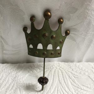 May include: A metal wall hook shaped like a crown, painted in a distressed green with bronze detailing. The hook features a dark brown, rounded knob. The crown design has a vintage, antique appearance.