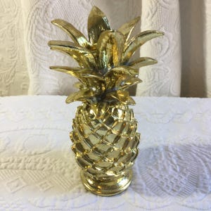 May include: A gold-coloured pineapple ornament. The pineapple has a textured surface and a detailed crown of leaves. The base is round and the ornament is standing upright.