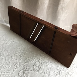 May include: A rectangular brown wooden mandoline slicer featuring a long, angled metal blade set into the center of the frame. The tool has a smooth, dark wood finish with two small metal fasteners visible along the top edge of the wooden body.