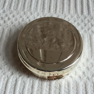 Puede incluir: Un compacto redondo plateado con una superficie de espejo. El compacto tiene un cierre de bisagra y un borde ligeramente elevado. La superficie del espejo tiene pequeñas manchas oscuras. El compacto está sobre una superficie blanca texturizada.