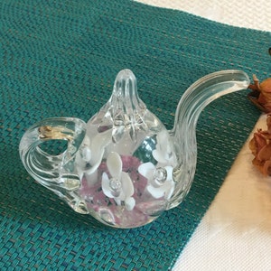 Könnte beinhalten: Eine klare Glasteekanne mit einem rosa-weißen Blumendesign im Inneren. Die Teekanne hat einen gebogenen Ausguss und einen Henkel.