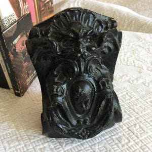 May include: Black and white decorative wall sculpture featuring a grotesque face and a heart-shaped design. The sculpture is made from a textured material and has a distressed look.