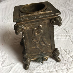 May include: A small, antique, bronze-colored inkwell with a square base and four decorative feet. The inkwell has a round, open top and a raised rim. The sides of the inkwell are decorated with a relief sculpture of a figure playing a musical instrument.
