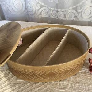 Puede incluir: Una caja ovalada de cuero marrón con tapa. La caja tiene tres compartimentos en el interior y un patrón decorativo en relieve en el exterior.