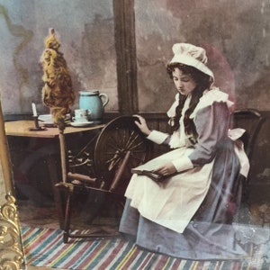 May include: A young woman in a white cap and blue dress sits by a spinning wheel, reading a book. She is in a room with a wooden table and a pitcher on it. The room has a striped rug on the floor.