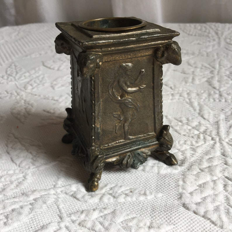 May include: A small, square, bronze-colored metal container with a decorative design. The container has a raised rim and four feet. The design features a figure of a woman in a flowing robe, holding a small object in her hand.