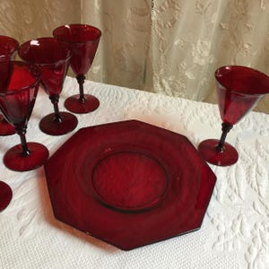 May include: A set of six ruby red glass wine glasses and a matching octagonal plate. The glasses have a stemmed base and a round bowl. The plate has a raised rim and a center well.