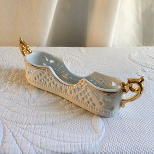 May include: White ceramic serving dish with gold trim and handles. The dish has a decorative cut-out design.
