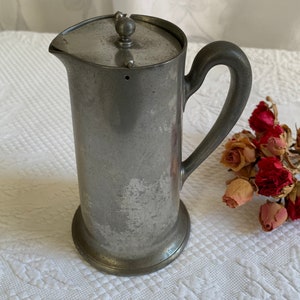 Puede incluir: Una jarra de peltre vintage con tapa y asa. La jarra tiene un aspecto desgastado y envejecido y está hecha de un metal plateado.