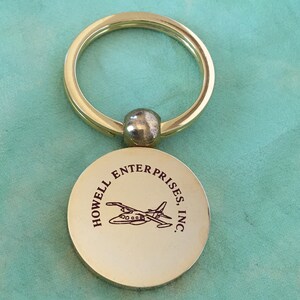 May include: A gold-colored metal keychain with a circular tag. The tag features the text "HOWELL ENTERPRISES, INC." around a black graphic of an airplane. The keychain ring is large and circular.