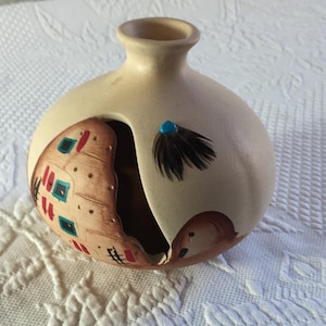 May include: A beige ceramic vase with a round body and a narrow neck. The vase features a cut-out section with painted details resembling a dwelling. A turquoise bead and feathers are attached to the exterior.