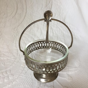 May include: Silver sugar bowl with a glass insert and a small spoon attached to the handle. The bowl has a decorative, openwork design.