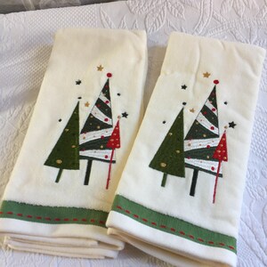 Puede incluir: Dos toallas de mano color blanco hueso con diseños bordados de árboles de Navidad en verde, blanco y rojo. Cada toalla tiene un borde verde en la parte inferior con una línea cosida en rojo. Los árboles incluyen pequeñas estrellas doradas y negras.