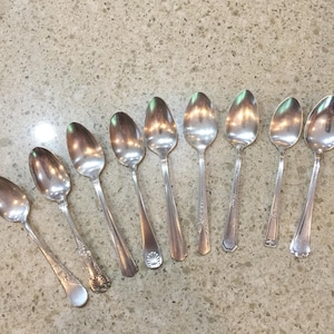 Puede incluir: Una colección de ocho cucharas de plata con diferentes diseños de mango. Las cucharas están dispuestas sobre una superficie clara y moteada. Las cucharas son reflectantes, mostrando la luz y el entorno.