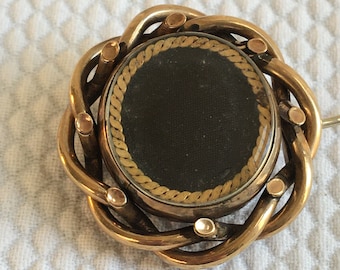May include: A gold-toned locket with a black fabric interior and a gold-coloured rope design around the edge. The locket is surrounded by a woven gold-toned metal frame.