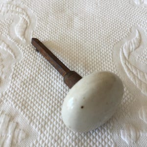 May include: A white ceramic door knob with a metal spindle. The knob is slightly oval shaped and has a small brown spot on the surface.