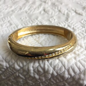 May include: A gold-toned bangle bracelet with a row of small, clear rhinestones. The bracelet has a smooth, polished finish and a hinged clasp. The background is a white, textured fabric.