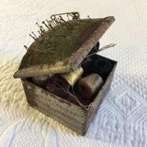 May include: An open, antique sewing box with a hinged lid. The lid has a green pin cushion with numerous pins and a safety pin. Inside the box are spools of thread, a thimble, and other sewing notions. The box is made of metal with a distressed finish.