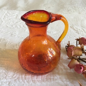May include: A small, orange glass pitcher with a red rim and a crackled glass finish. The pitcher has a round body and a curved handle.