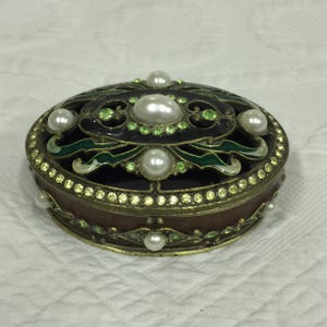 May include: An oval-shaped trinket box with a black and green enamel design. The box is decorated with faux pearls and rhinestones.