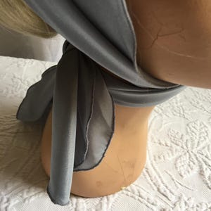 May include: A light gray scarf draped around a mannequin's neck. The scarf is made of a soft, flowing fabric and has a delicate, slightly ruffled edge. The mannequin has blonde hair and is positioned on a white patterned surface.