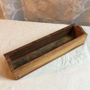 May include: A rectangular wooden tray with a natural finish. The tray has a long, narrow design with a dark brown interior and lighter brown exterior. The wood grain is visible, giving it a rustic appearance. The tray is empty.