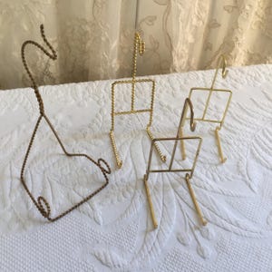 以下が含まれることがあります： さまざまなデザインの、4つの小さな装飾用金属製ディスプレイスタンド。1つは茶色、その他は金色です。スタンドはワイヤー製で、小さなアイテムを保持するように設計されています。スタンドは白い表面にあります。