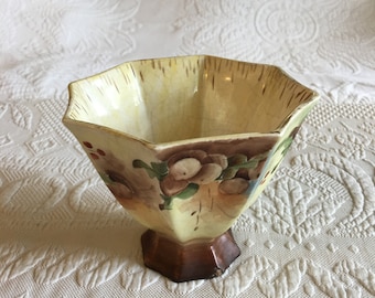May include: A small, octagonal, yellow ceramic bowl with a brown base. The bowl is decorated with a floral pattern in shades of brown, green, and red.