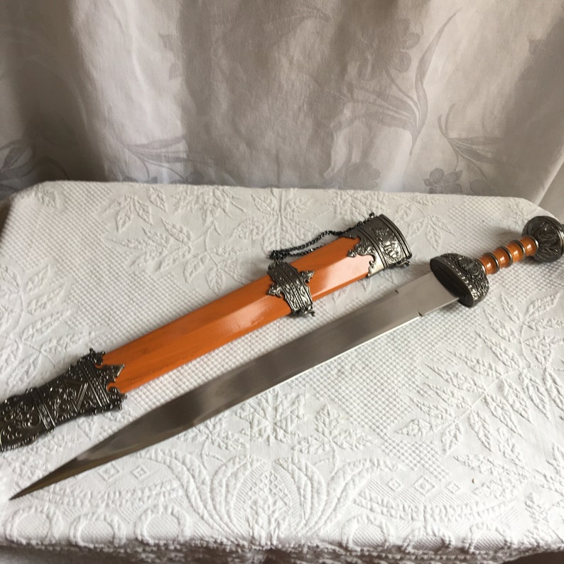 Puede incluir: Una espada plateada y naranja con un mango y una vaina decorativos. La espada tiene una hoja larga y afilada y una empu&ntilde;adura detallada. La vaina es de madera y tiene una punta de metal.