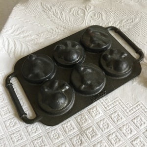 May include: A cast iron baking pan with six round moulds. The moulds are shaped like animal faces. The pan has a black finish and a single handle.