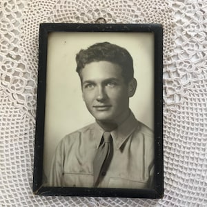 May include: Black and white framed portrait of a man in a button-down shirt and tie. The portrait is in a black rectangular frame with a small metal loop at the top for hanging. The photograph appears to be vintage.