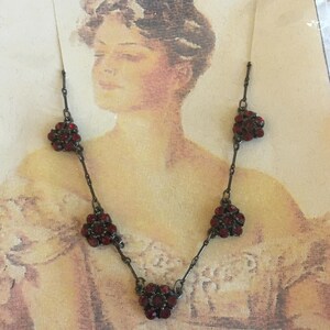 May include: A vintage necklace featuring dark red floral clusters. The necklace has a dark metal chain and is set against a vintage-style background. The floral clusters are made of small, dark red stones.