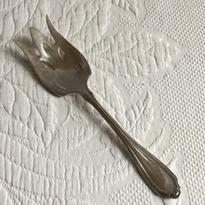 Op de afbeelding: Een zilveren serveersleutel met een decoratief handvat en een gegolfde rand. De vork heeft een uniek ontwerp met een uitgesneden patroon in de tanden.
