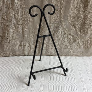 May include: A black metal easel with a three-legged design. The easel has a decorative top with two curved swirls. The easel is designed to hold a small picture frame or decorative item. The easel is made of metal.