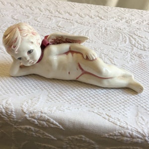 May include: A white ceramic cherub figurine with pink accents, lying on its side with its head resting on its hand. The cherub has wings and a pink ribbon around its neck.