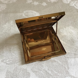 May include: A gold-toned, rectangular compact mirror, open to reveal a mirror and a compartment. The compact has a hinged lid and a clasp. The interior is visible. The item is on a white, patterned surface.